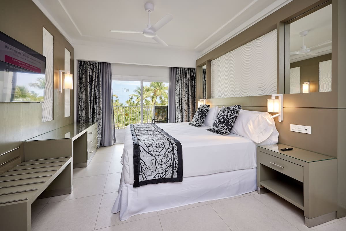 A modern hotel room with a large bed, bedside tables, a ceiling fan, and a view of palm trees.