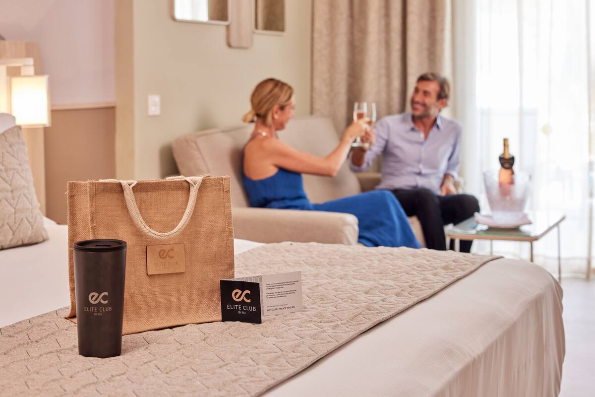 A hotel room with a bed, a bedside table with a bottle, and a bag labeled "ELITE CLUB".