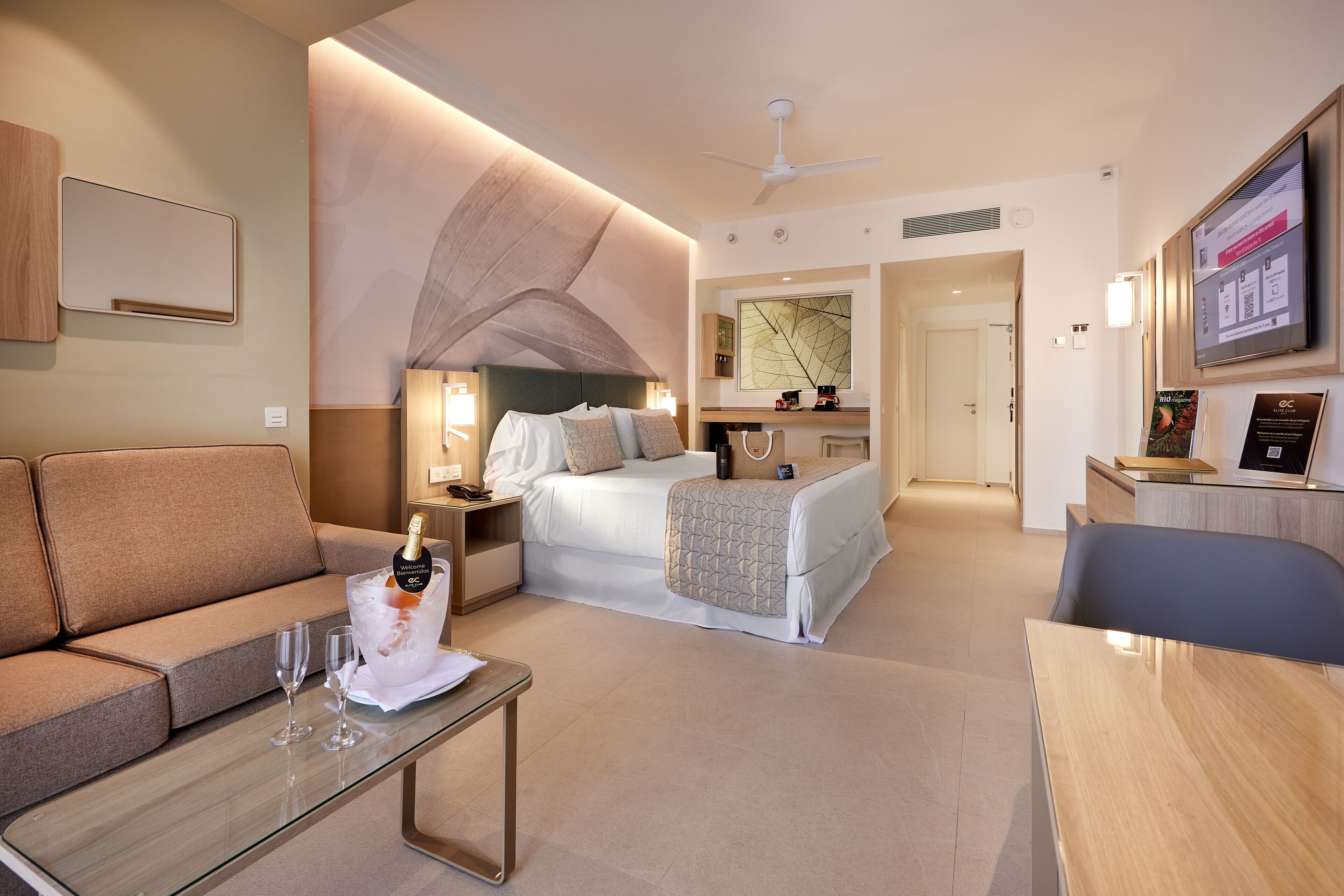 Habitación de hotel con cama, sofá, mesa de centro con vasos, televisión y minibar.