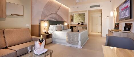 Habitación de hotel con cama, sofá, mesa de centro con vasos, televisión y minibar.