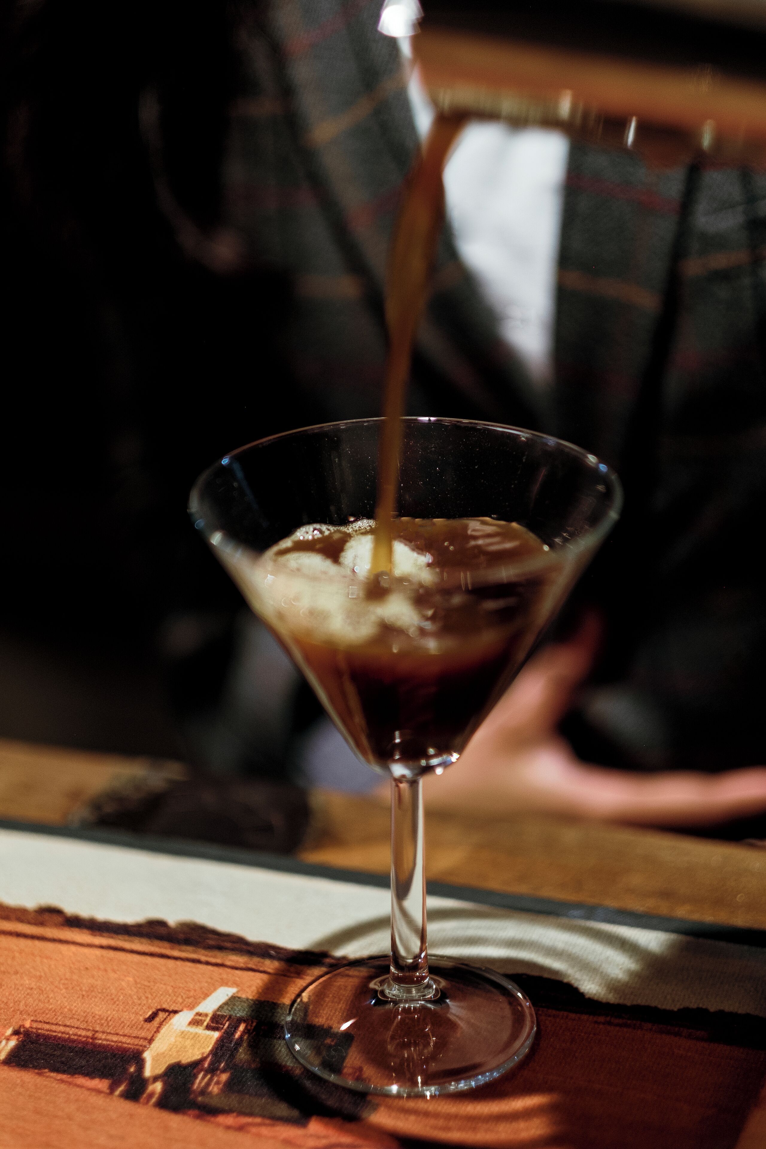 A martini glass with a dark liquid being poured into it, placed on a table with a plaid cloth.