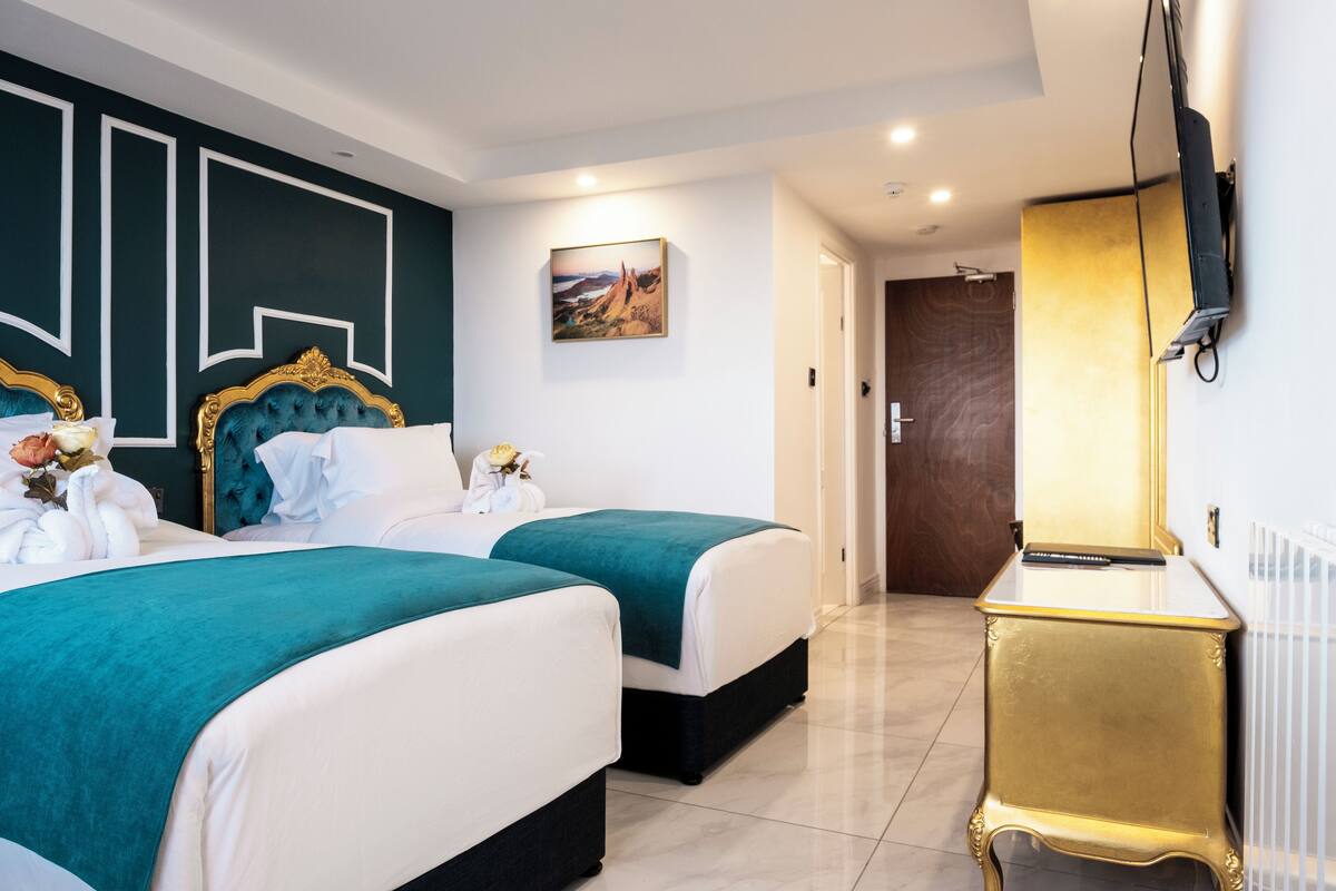 Habitación de hotel con dos camas, una mesita de noche con marco dorado, un televisor de pantalla plana y un cuadro en la pared.
