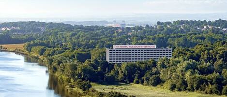 Un grand bâtiment sur lequel on peut lire l’enseigne « MGM Muthu Glassgow River Hotel », entouré d’une végétation luxuriante et bordé par une rivière.
