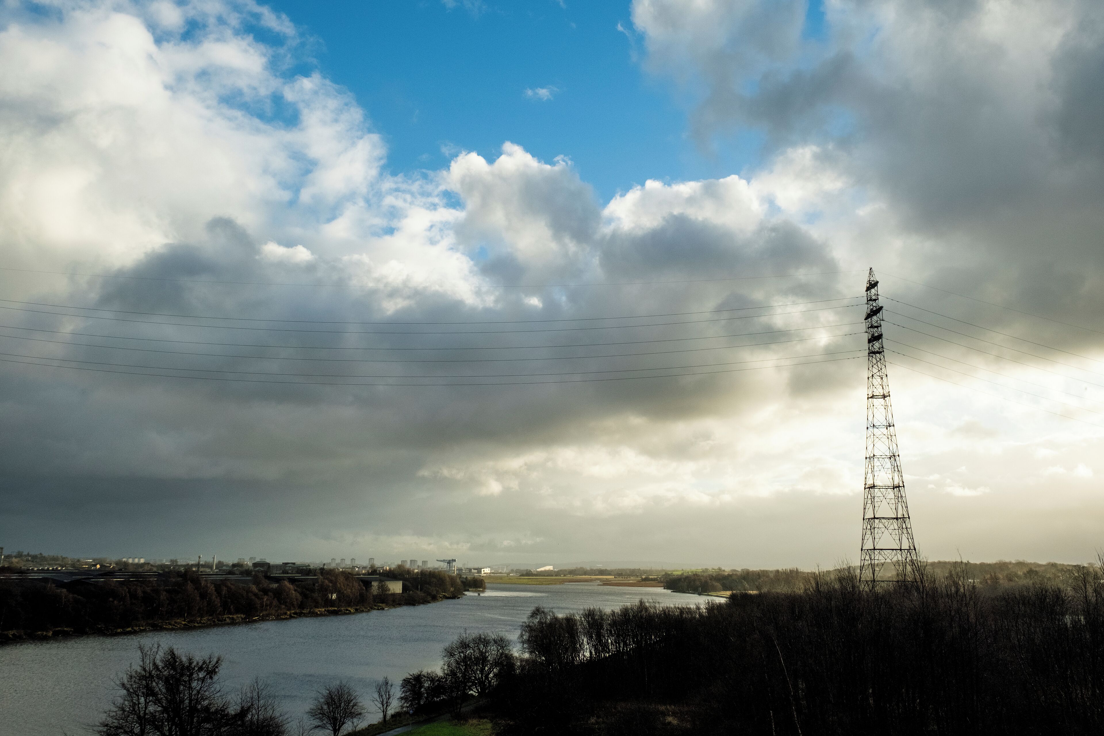 A river with a power line tower in the foreground and a cloudy sky.