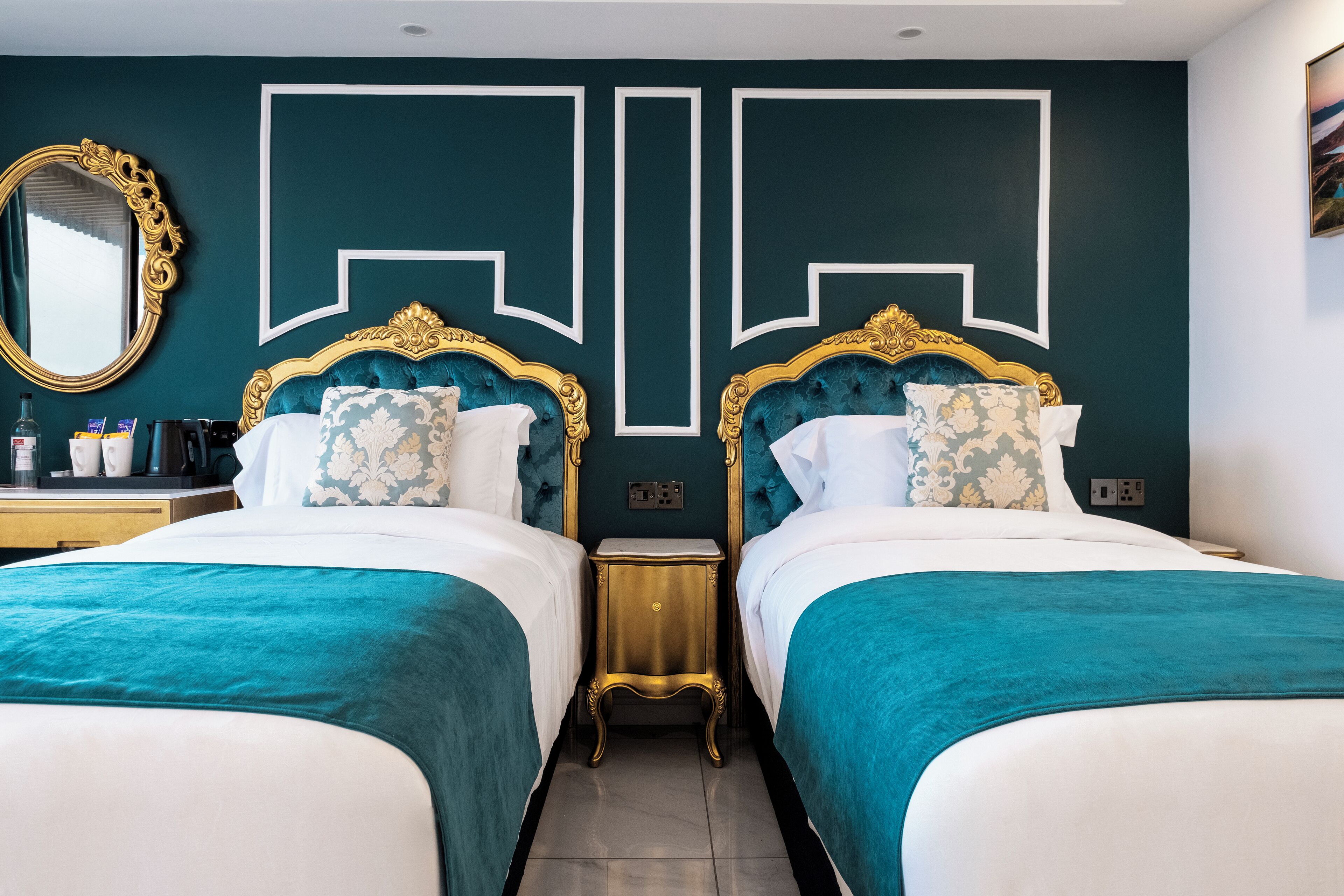 A hotel room with two beds, each with a teal bedspread and white linens, flanked by ornate gold headboards. A large mirror with a gold frame is mounted on the wall above the beds.