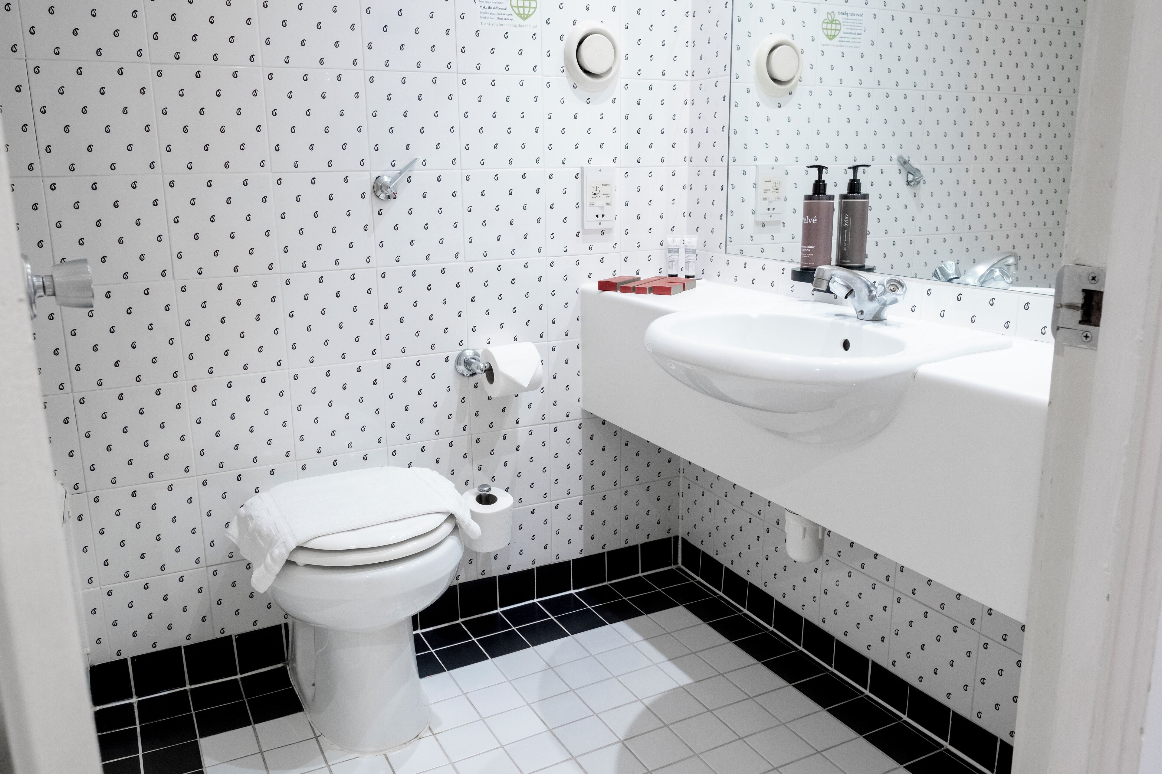 Ein Badezimmer mit Toilette, Waschbecken und Spiegel. Die Wände zeigen ein Muster aus kleinen Buchstaben und der Boden ist in Schwarz und Weiß gefliest.