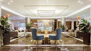 Een lobby met een marmeren vloer, een kroonluchter, twee blauwe fauteuils, een bank en een salontafel.