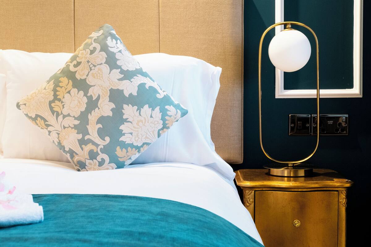 Una cama bien hecha con una colcha color turquesa, un cojín floral y una mesita de noche dorada con lámpara.