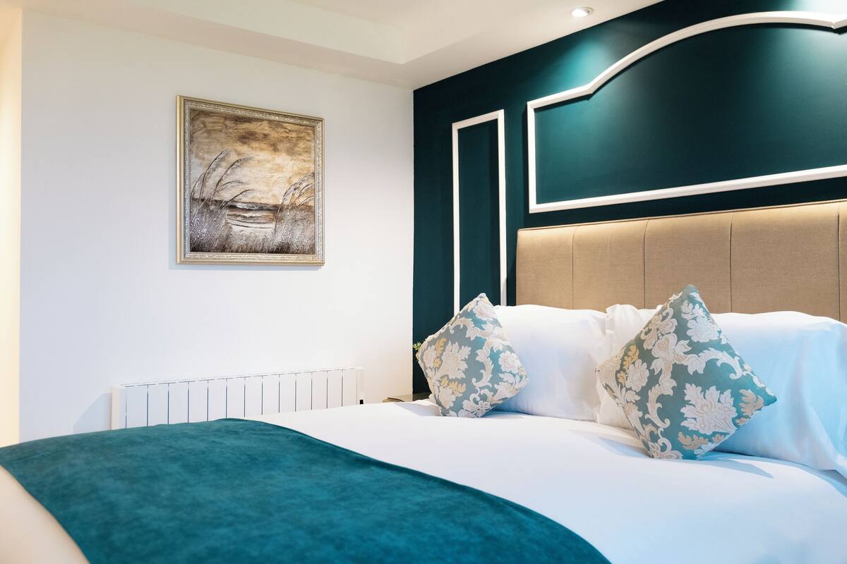 Un dormitorio moderno con una cama grande, dos almohadones decorativos, un cabecero con iluminación integrada y una obra de arte enmarcada en la pared.