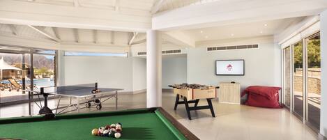A pool table with billiard balls, a foosball table, and a ping pong table in a spacious indoor area with large windows.