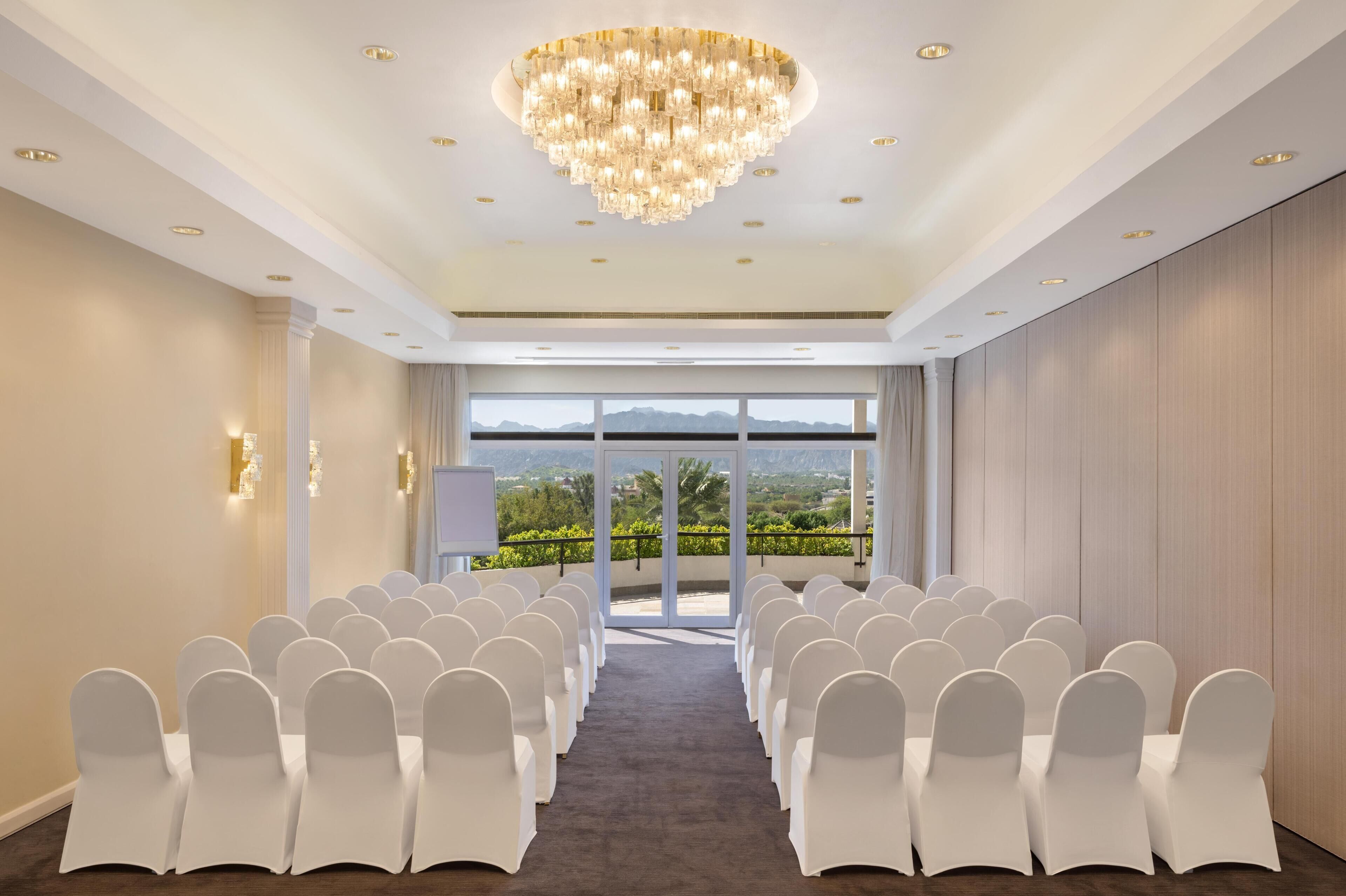 Un salón de conferencias con filas de sillas blancas, una lámpara de araña grande y vistas a través de puertas de cristal.