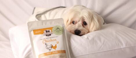 A dog lying on a white bed with a bag of dog food labeled "Be Our Child Chicken" in front of it.