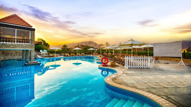Une piscine entourée d’une terrasse en pierre, avec des chaises longues, des parasols et une vue sur les montagnes.