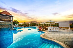 A swimming pool with a stone deck, lounge chairs, umbrellas, and a view of mountains. - JA Hatta Fort Hotel (Hatta)
