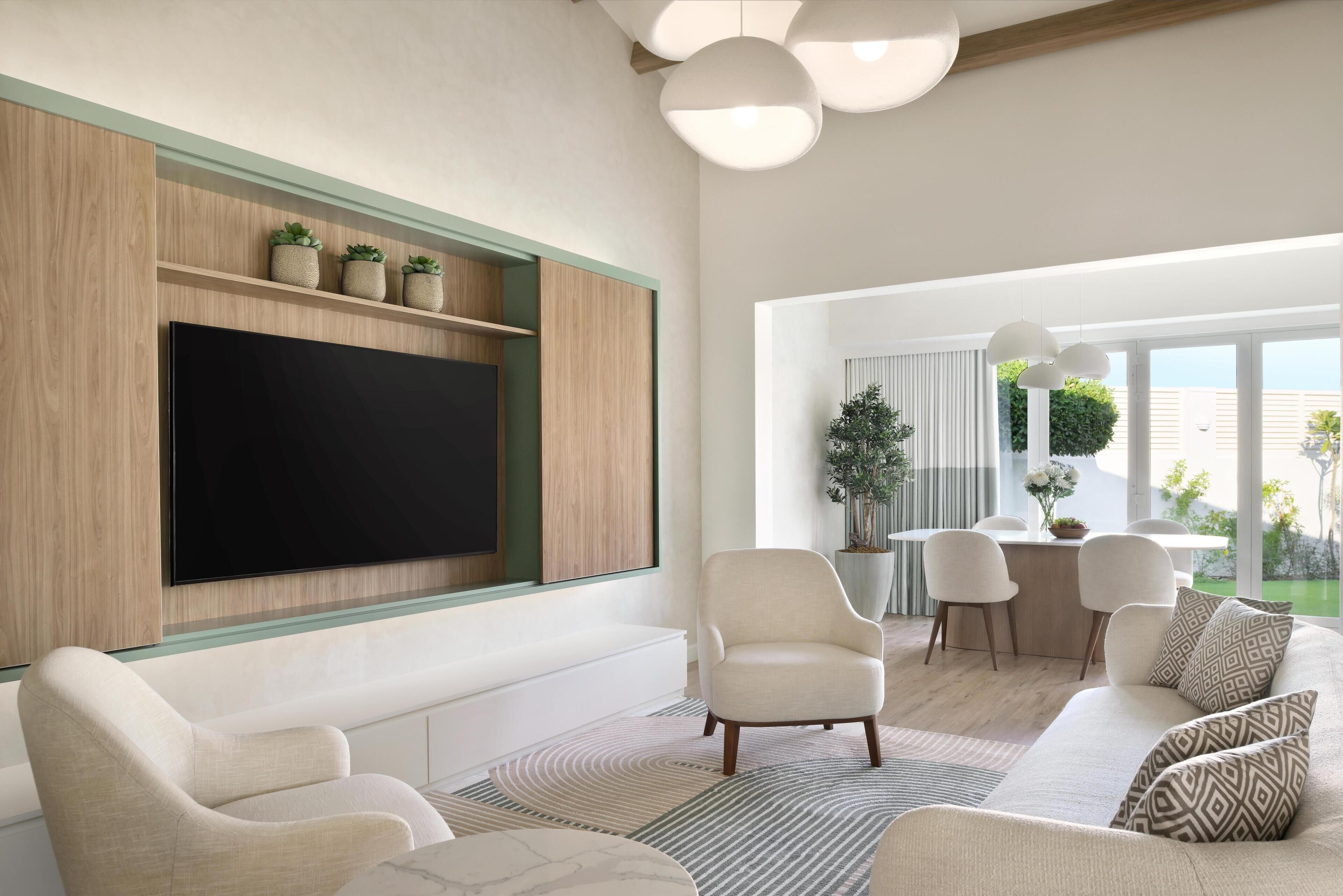 A modern living room with a large flat-screen TV, a sofa, armchairs, a dining area with a table and chairs, and a view of the outdoors.
