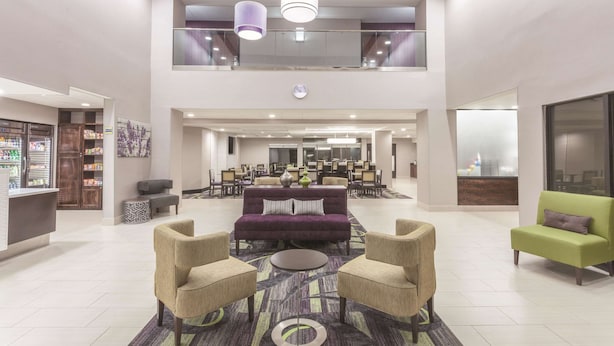Un hall d’entrée spacieux avec un canapé violet, des fauteuils beiges et un canapé vert. On y trouve un comptoir d’accueil, une horloge et une grande fenêtre.