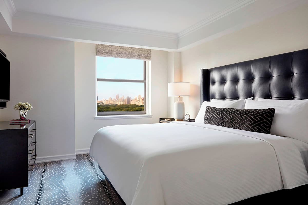 Habitación de hotel con una cama grande, una mesita de noche con lámpara, un aparador y una ventana con vistas a la ciudad.