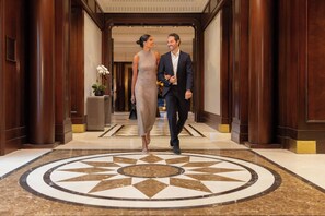 A man and a woman walking through a lobby with a patterned floor, wooden columns, and a potted plant. - JW Marriott Essex House New York (New York)