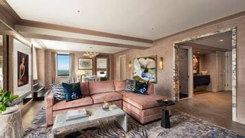 A spacious living room with a large sectional sofa, a coffee table, and a patterned rug. There is a large painting on the wall and a view of the outdoors through a window.