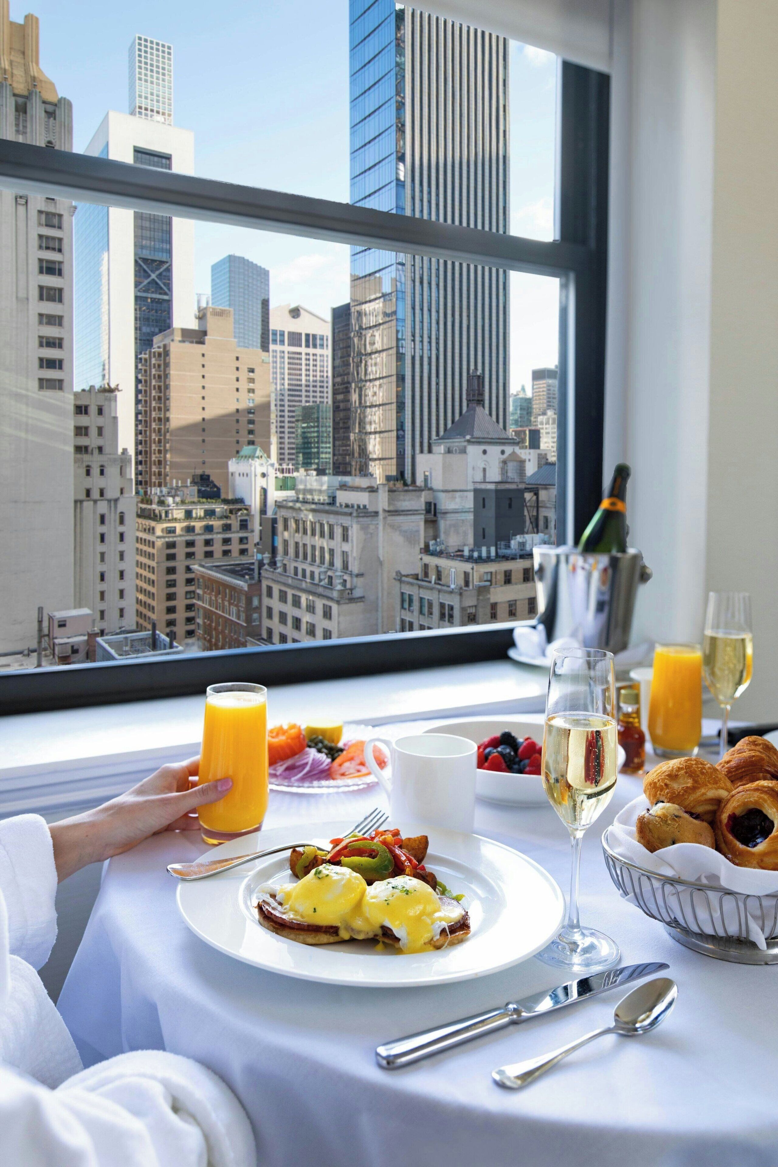 A breakfast table with a city view, including a bottle of champagne, a glass of orange juice, and croissants.