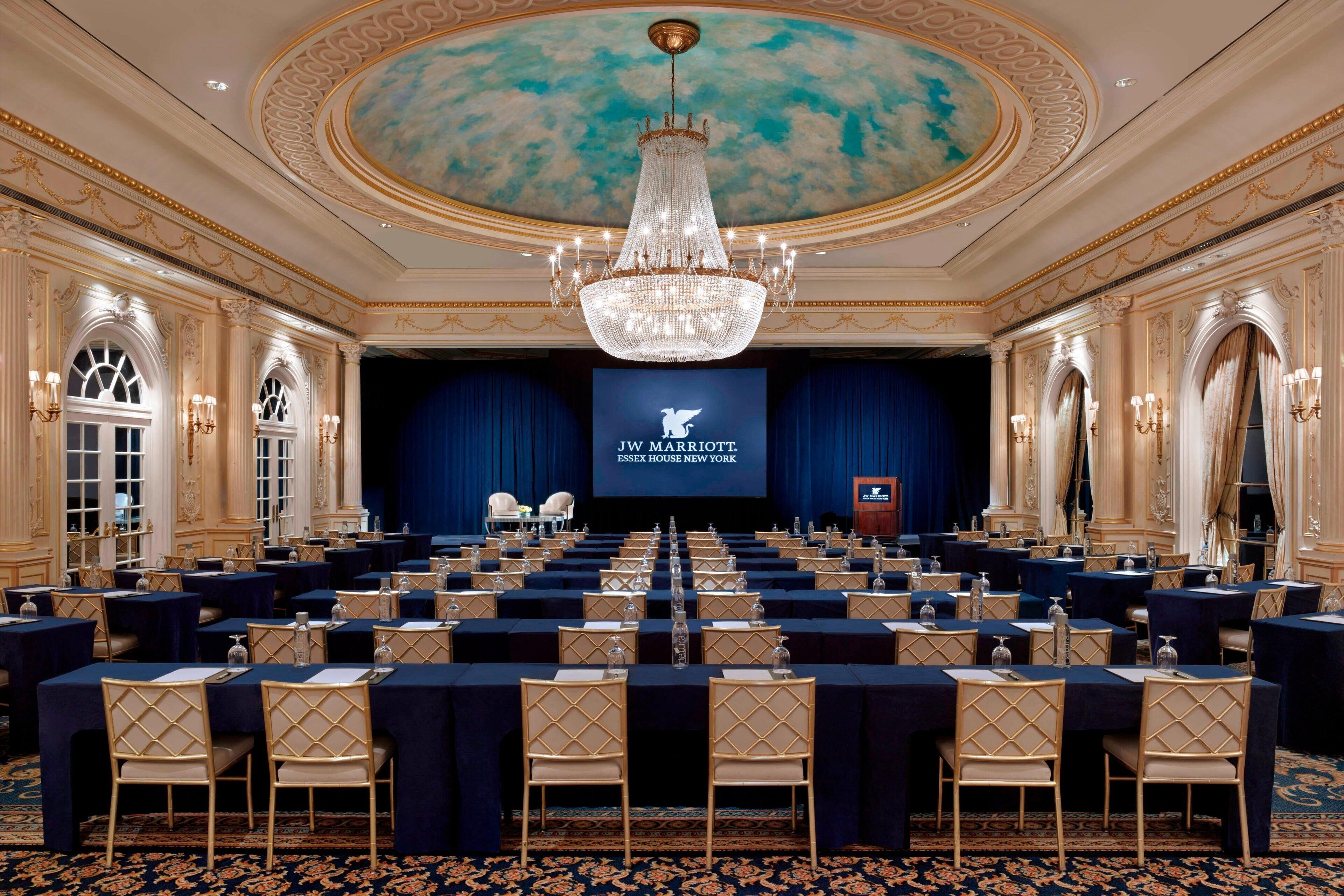 A conference room with rows of tables and chairs, a large chandelier, and a screen displaying the JW Marriott logo.