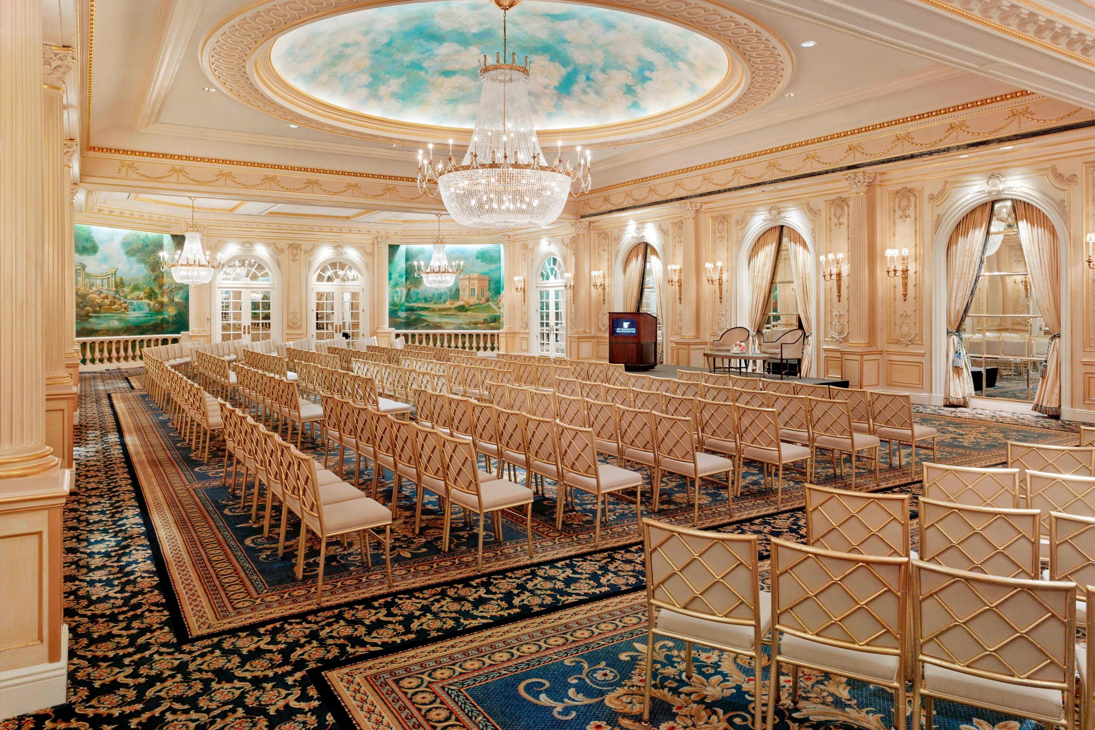 Un gran salón de baile con filas de sillas, una lámpara de araña imponente y pinturas murales ornamentales.