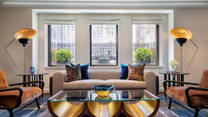 Una sala moderna con un sofá, dos sillones, una mesa de centro de vidrio y dos lámparas de pie. Se ven grandes ventanales con vistas a edificios y zonas verdes.