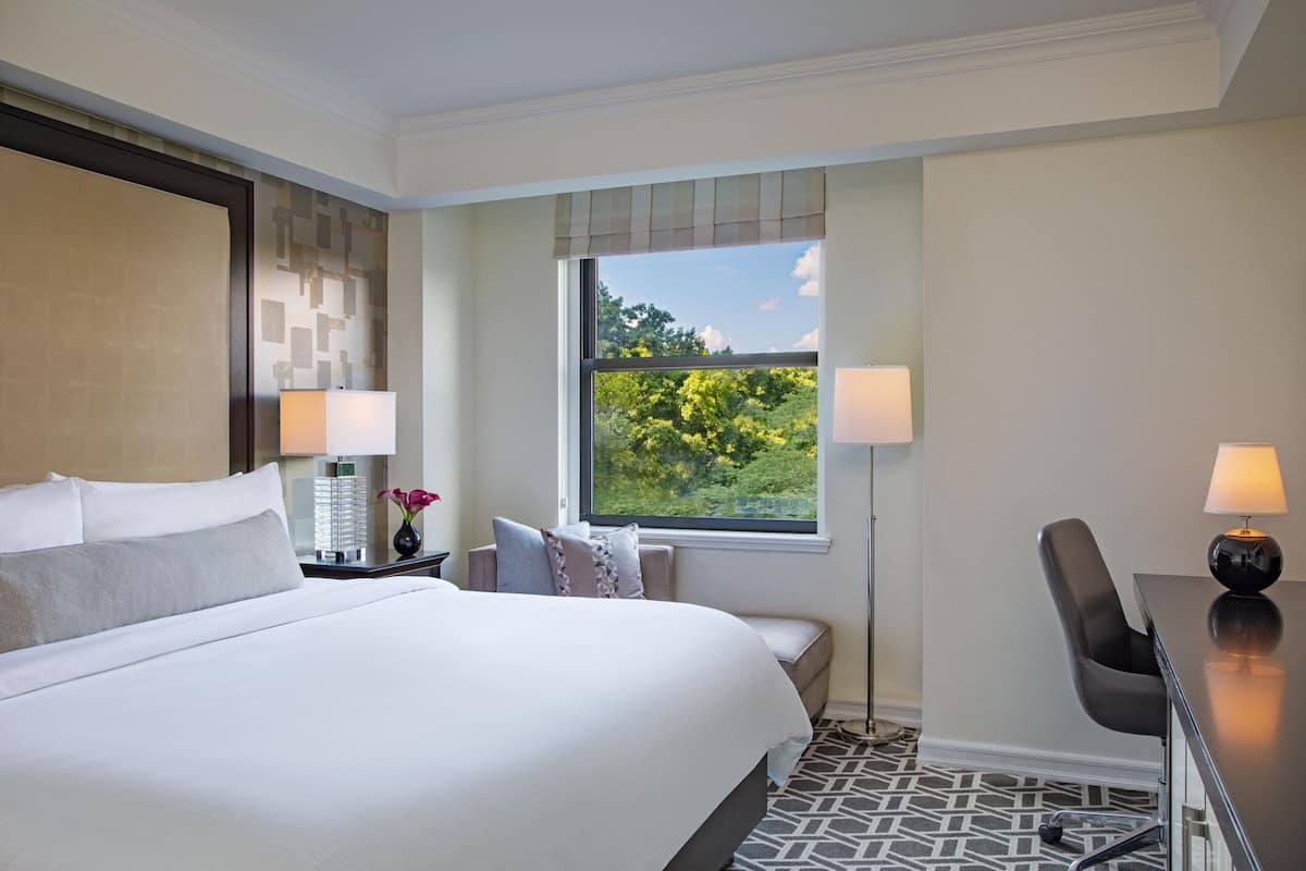 Habitación de hotel con una cama grande, dos lámparas de noche, un escritorio con silla y una ventana con vistas a la vegetación.