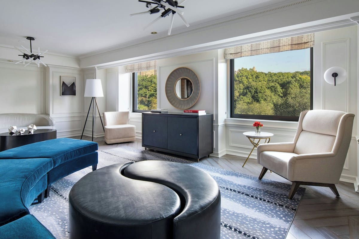 Una sala de estar moderna con un sofá azul, un otoman negro, un sillón beige y un mueble azul oscuro, con un ventanal que ofrece vistas a árboles.