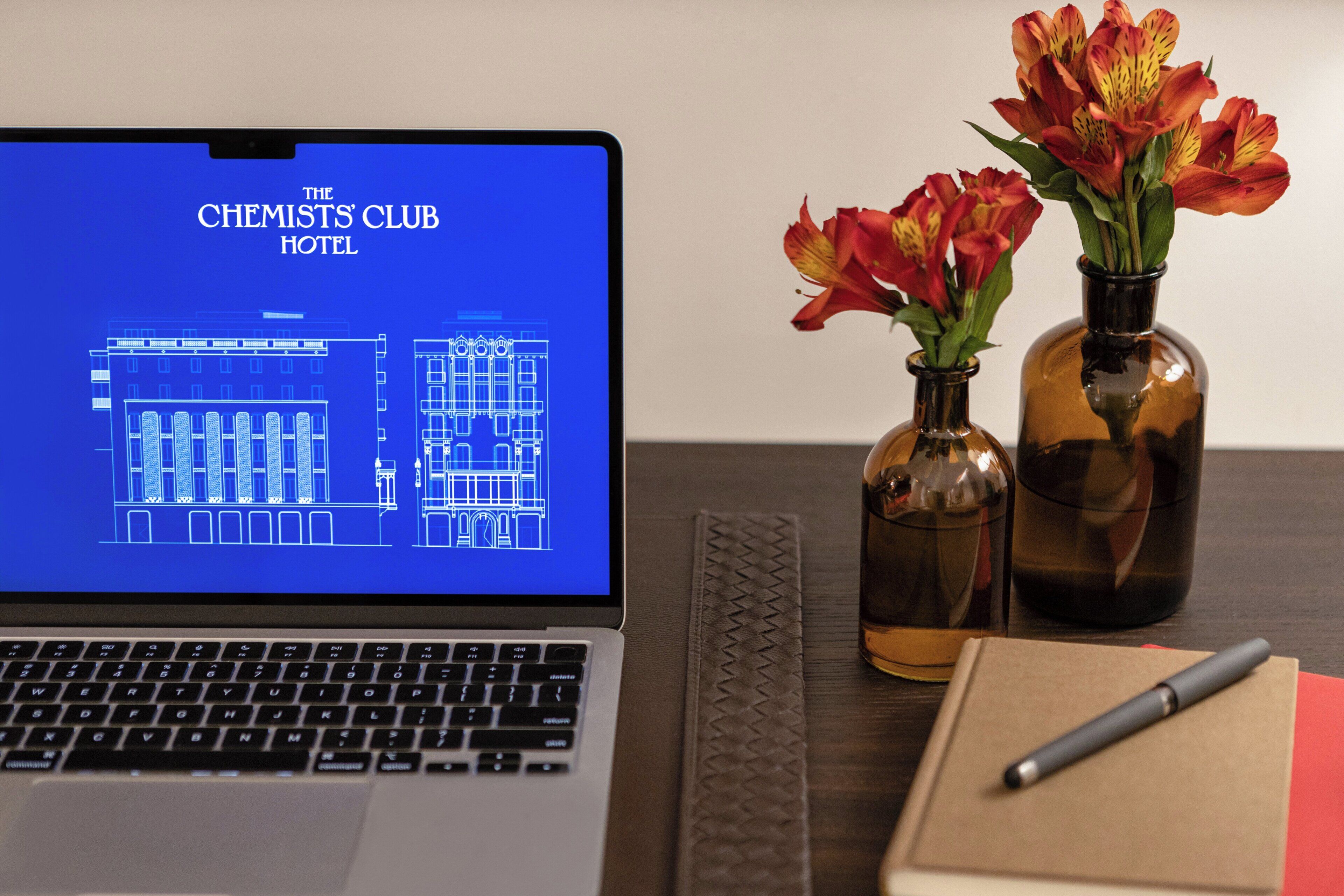 A laptop displaying a hotel blueprint, two vases with flowers, a notebook, and a pen on a table.
