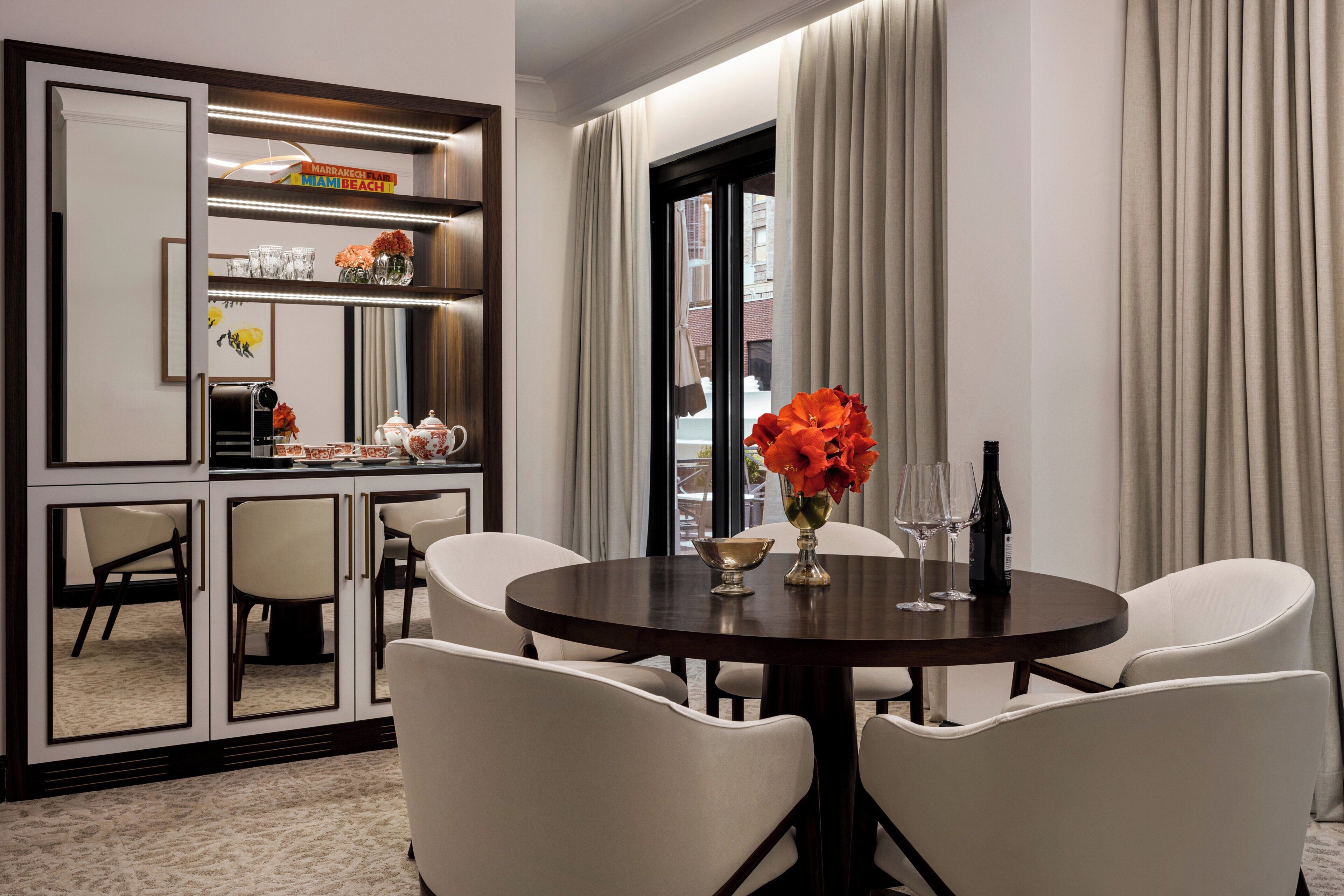 Quarto de hotel com mesa de jantar redonda, quatro cadeiras, espaço para café e uma janela grande com cortinas, além de um espelho refletindo o ambiente.