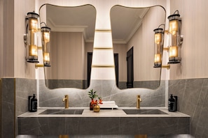 A bathroom with a large mirror, two wall-mounted light fixtures, a gold faucet, and a marble countertop. - Eurostars The Chemists’ Club Hotel (New York)