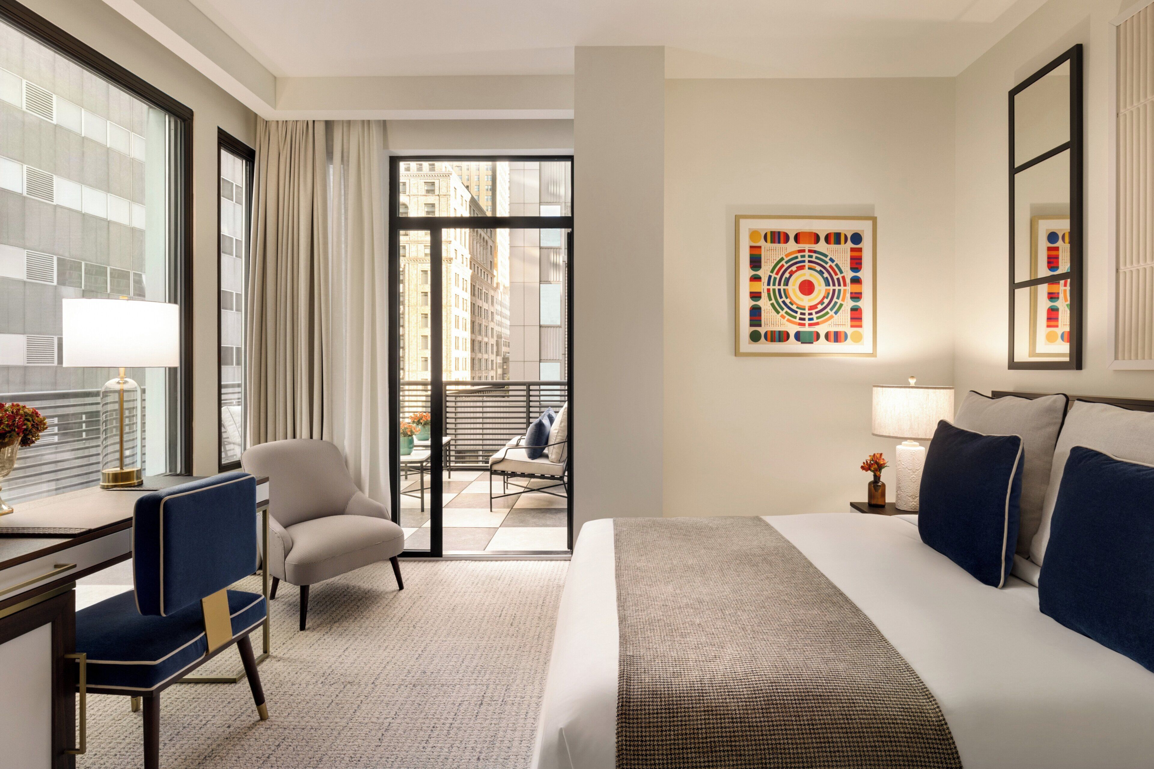 A hotel room with a bed, a desk, a chair, a balcony with a view of buildings, and a wall with a colorful abstract painting.