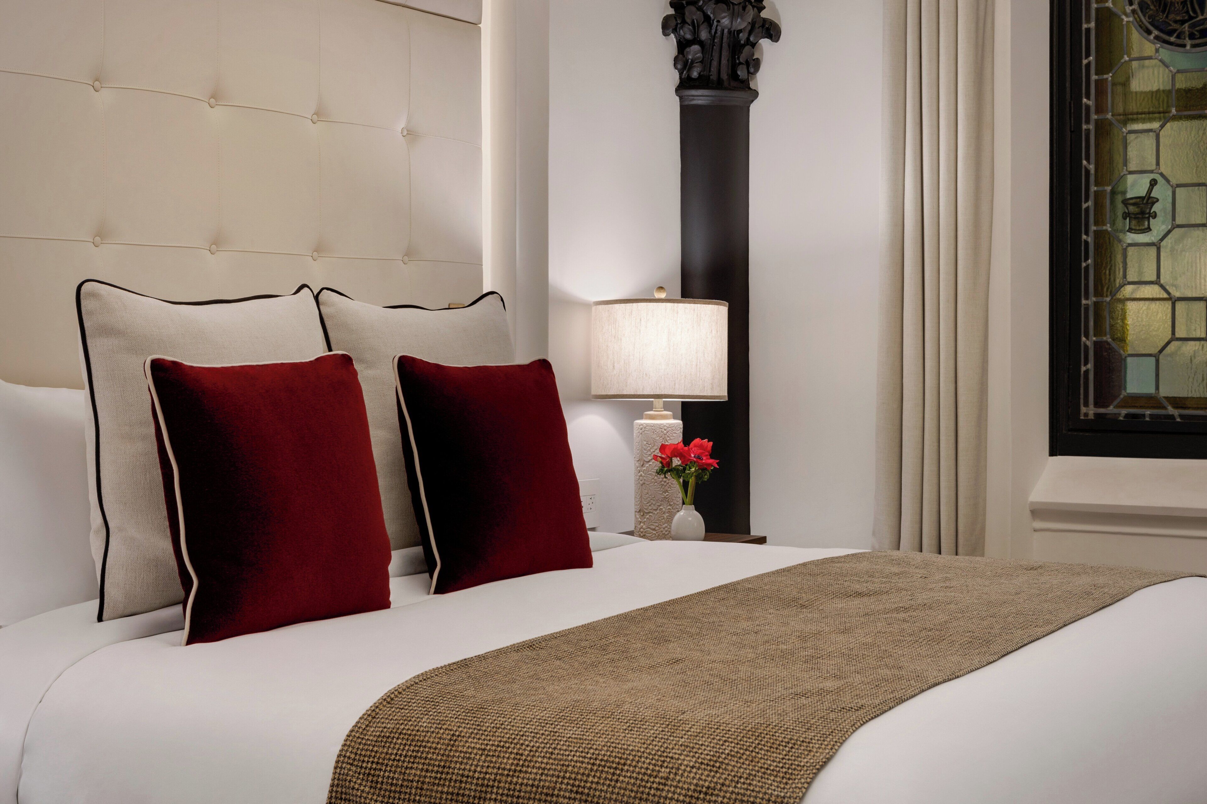A neatly made bed with a white headboard, two red pillows, a beige throw blanket, and a nightstand with a lamp and a vase of flowers.