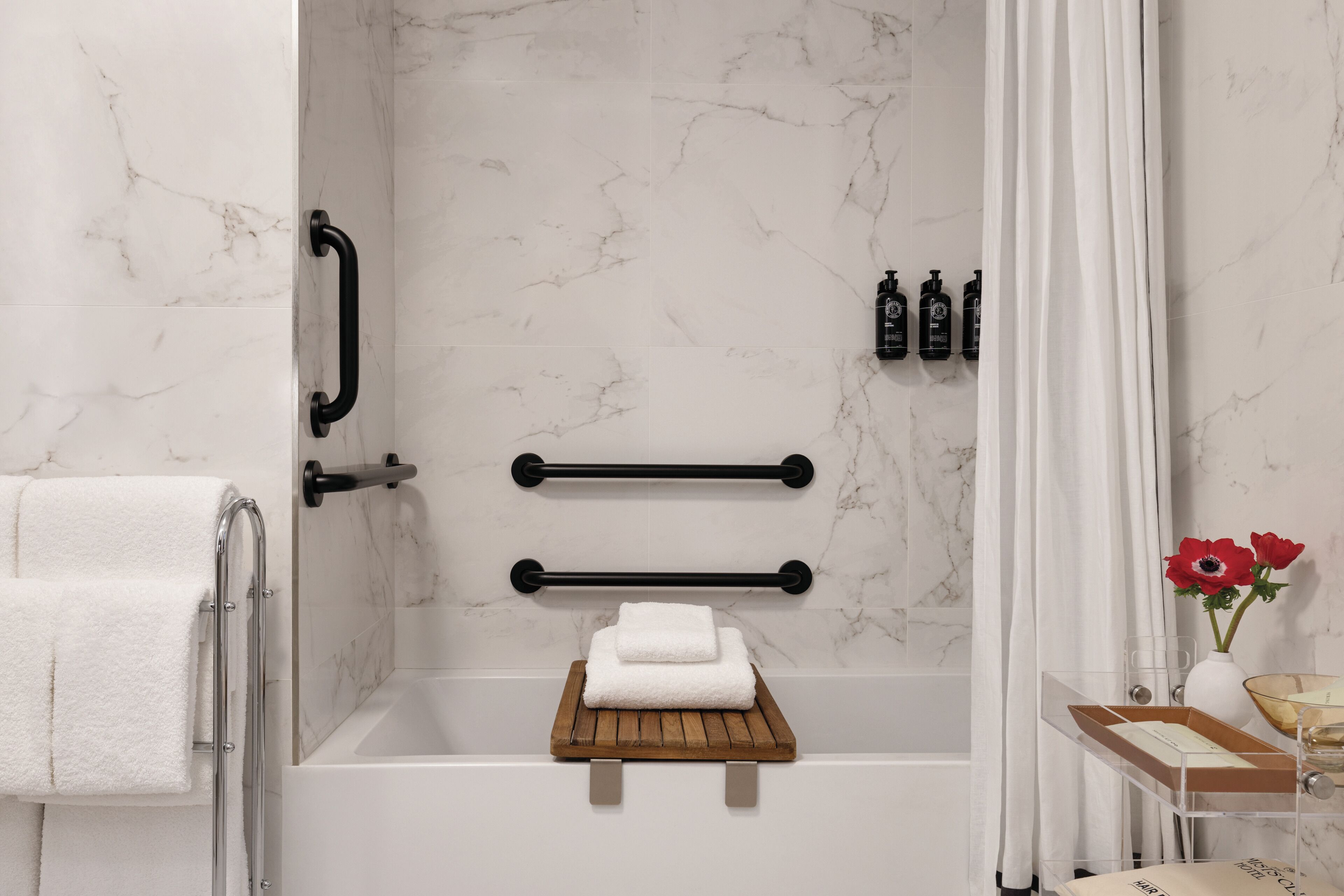 Een moderne badkamer met een badkuip, handgrepen, een houten plank met een handdoek en toiletartikelen aan de muur.