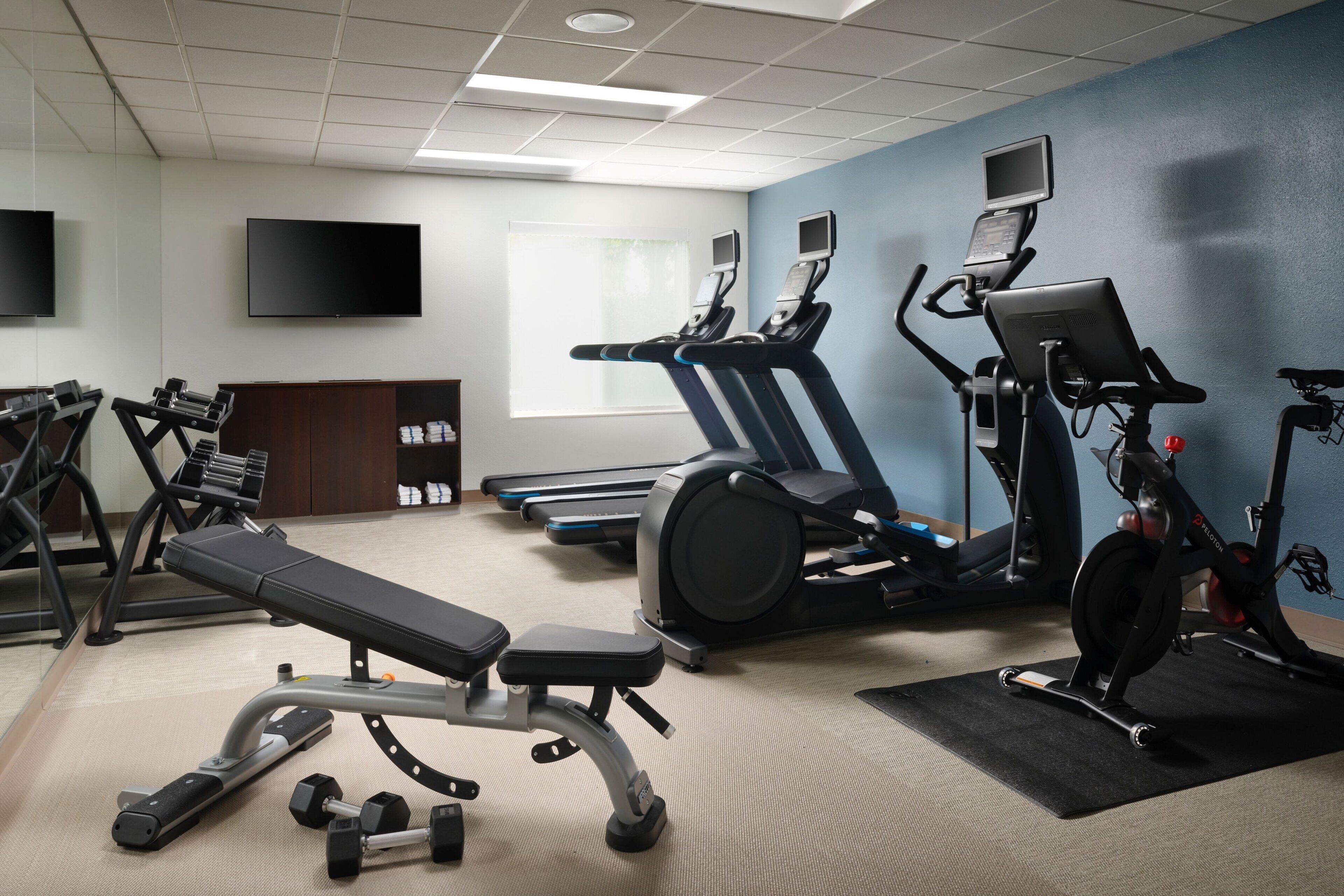 Ein Fitnessraum mit Trainingsgeräten, einer Bank, Kurzhanteln und einem Fernseher an der Wand.