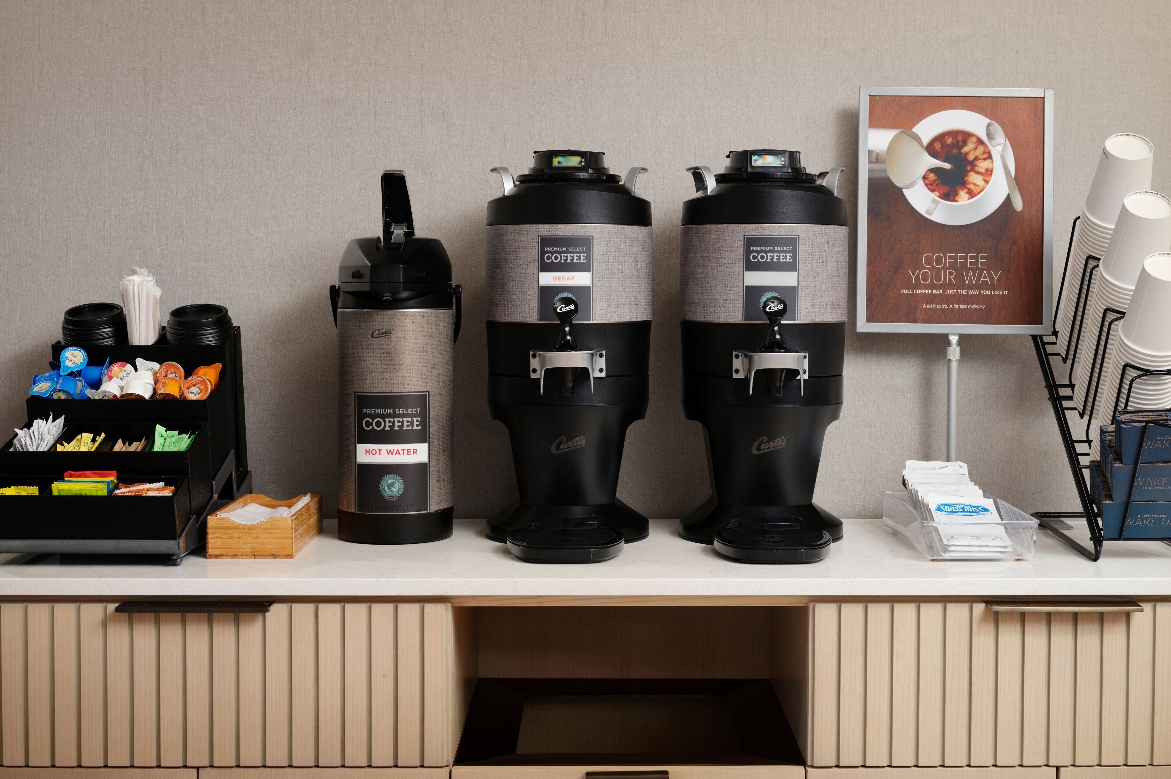 Een koffiehoek met drie koffieautomaten, een koffiekop en een poster.