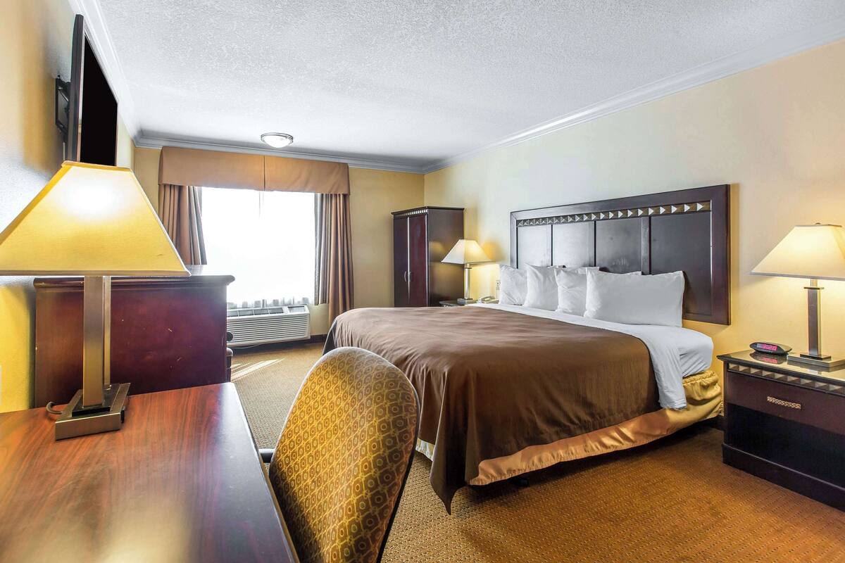 A hotel room with a large bed, two bedside tables with lamps, a wooden desk, a chair, and a wardrobe.