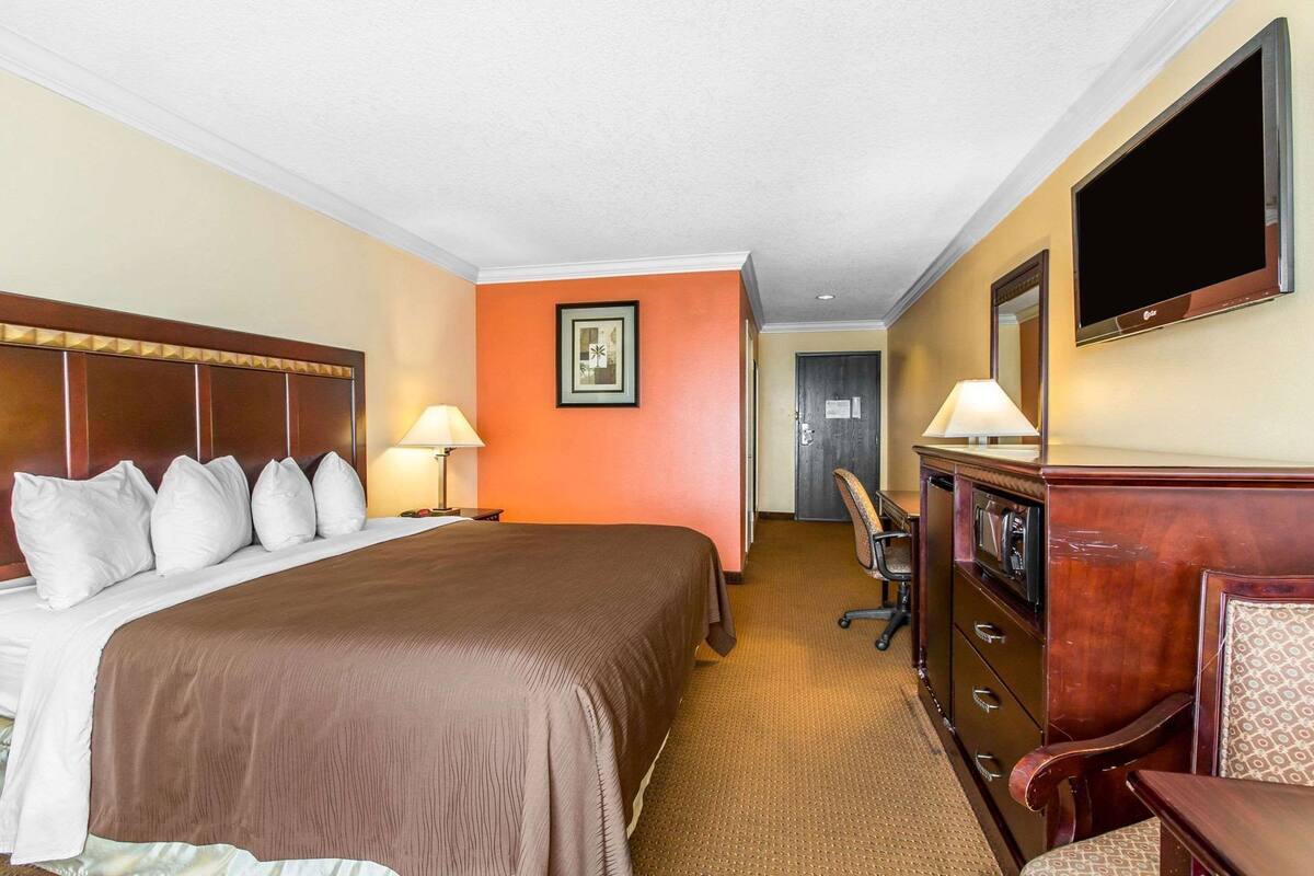 A hotel room with a bed, bedside lamps, a desk, a chair, a TV, and a door leading to another room.