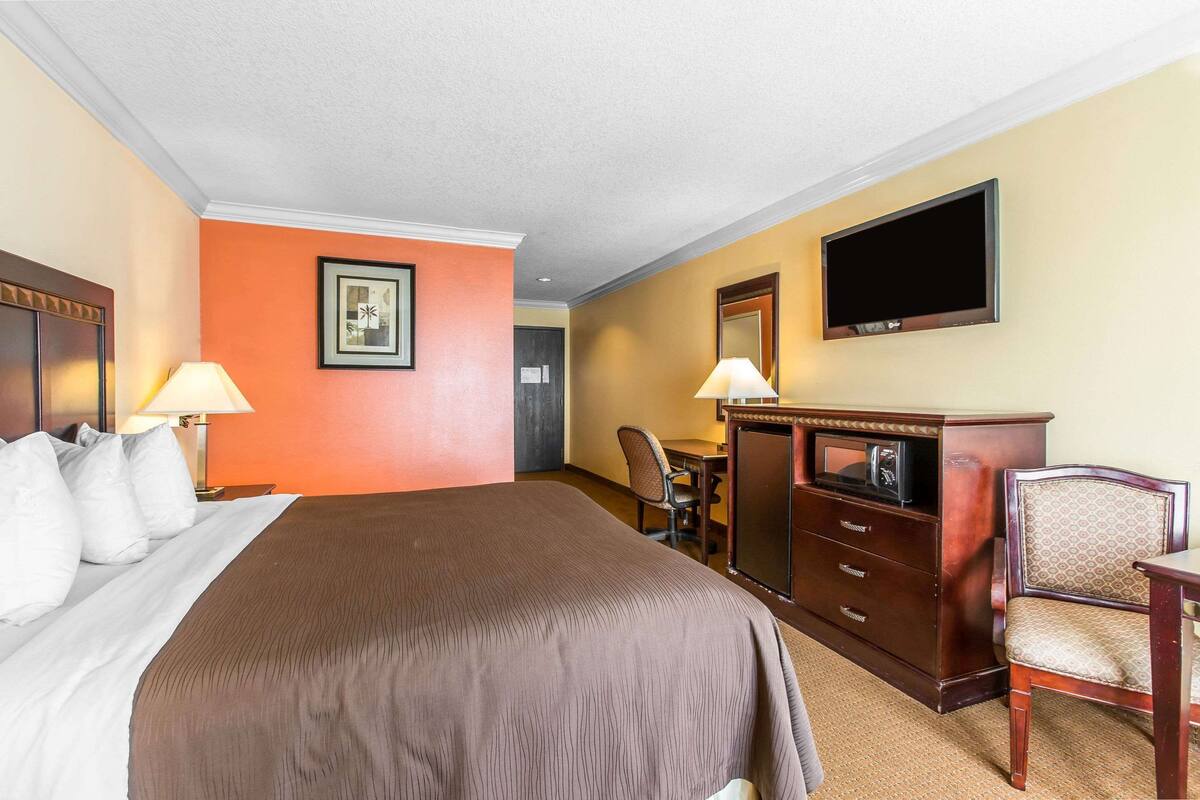 A hotel room with a large bed, two bedside lamps, a desk with a chair, a dresser, and a flat-screen TV mounted on the wall.