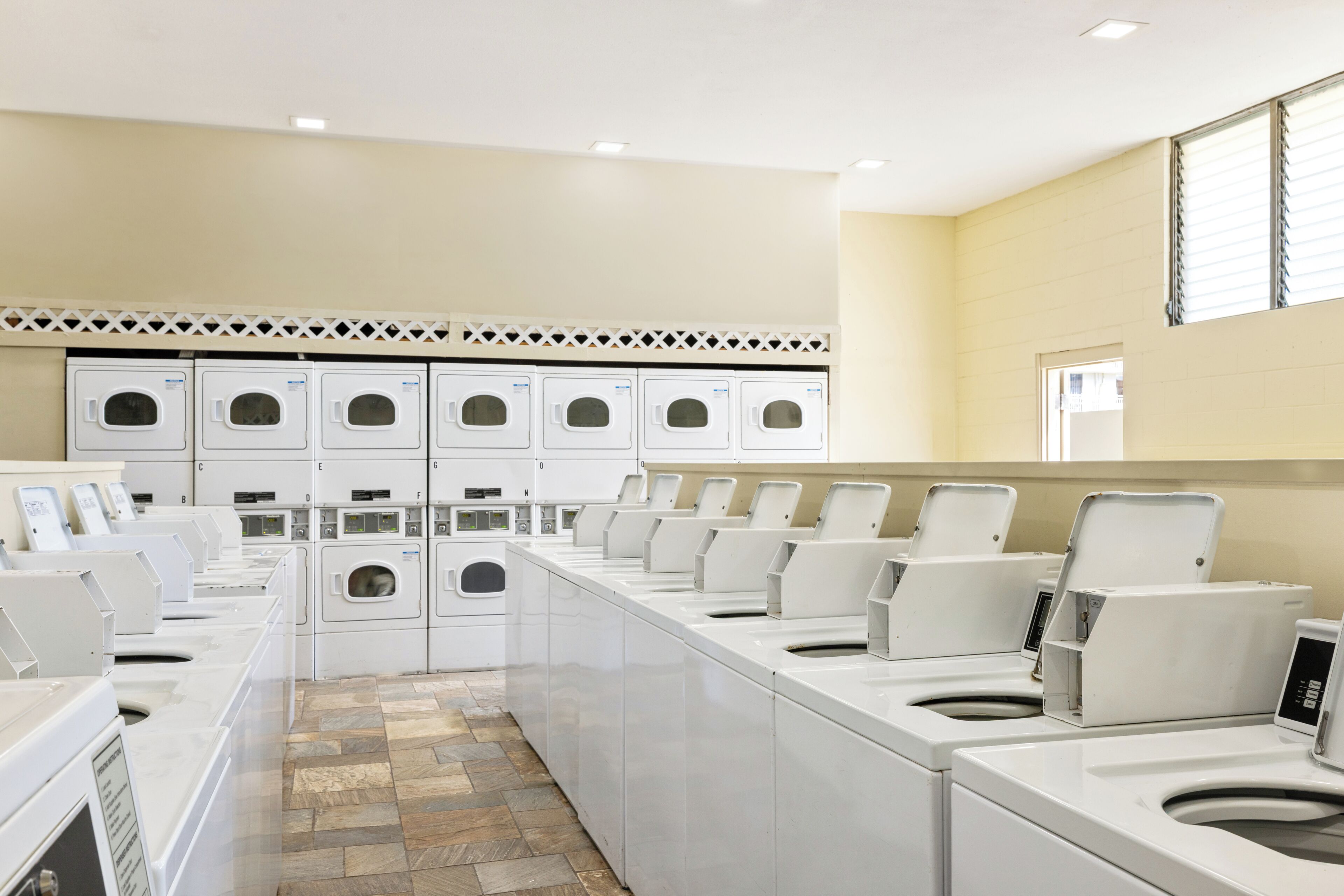 A laundry facility with multiple washing machines and dryers, a tiled floor, and a plain wall with a door.