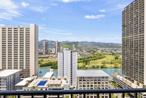 Un edificio de gran altura con varios pisos, un área de piscina con sombrillas y vistas a un paisaje urbano con montañas al fondo.
