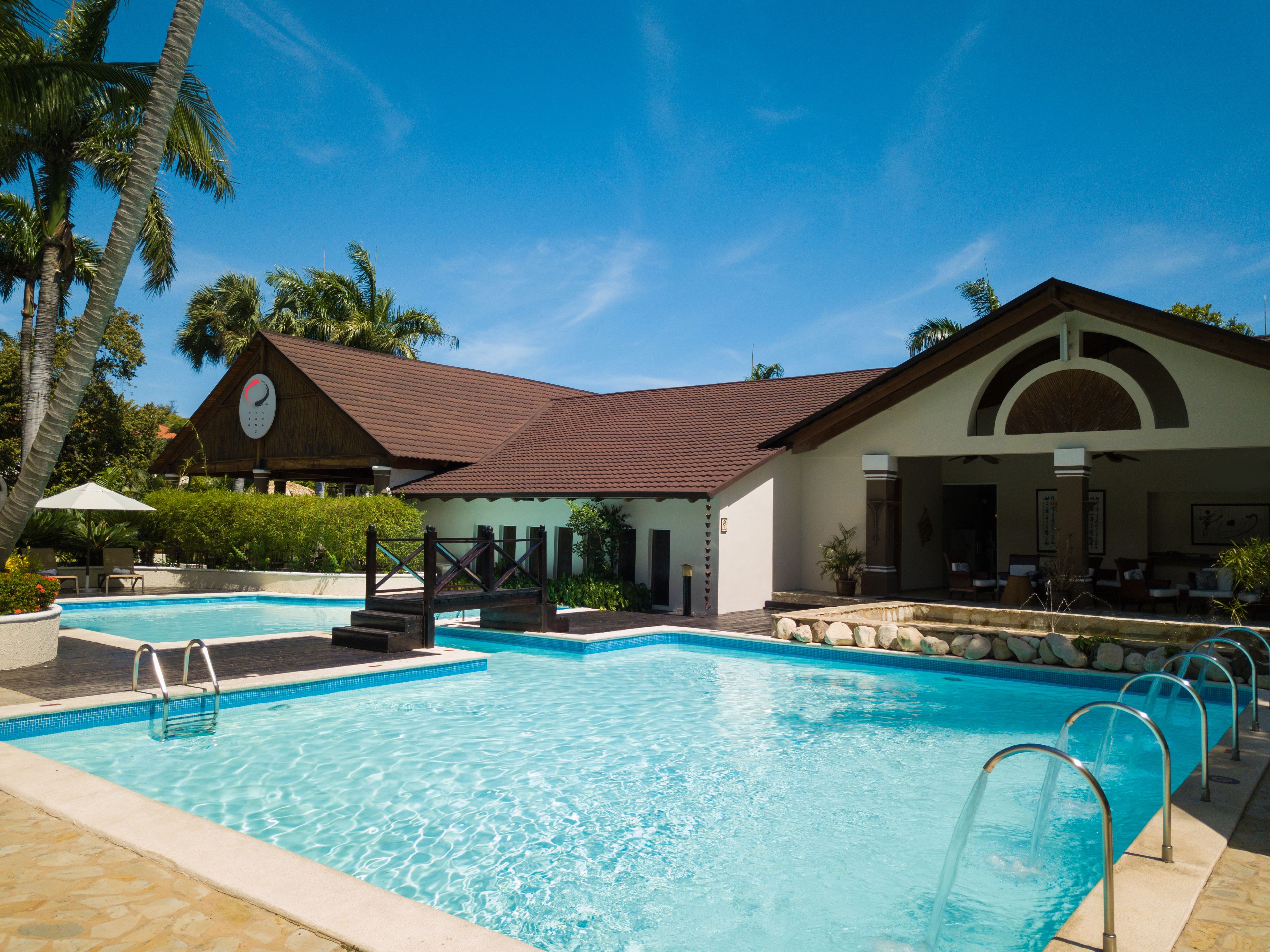 A swimming pool with a wooden deck, a covered seating area, and a building with a clock.