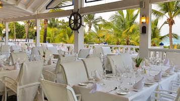 A restaurant with tables set for dining, overlooking a beach with palm trees and the ocean.
