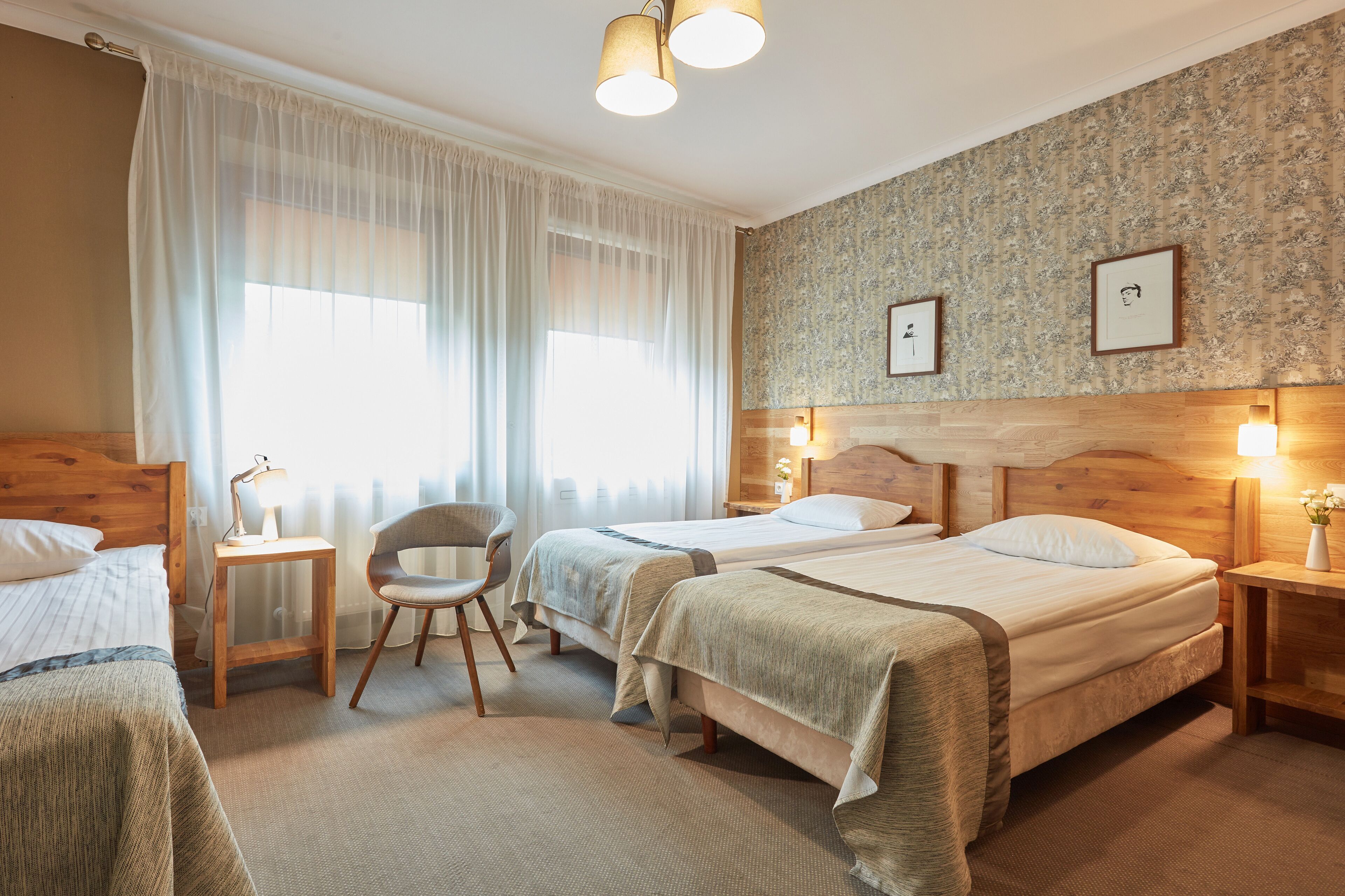 A hotel room with two beds, a chair, a nightstand with a lamp, and a wall with framed pictures.
