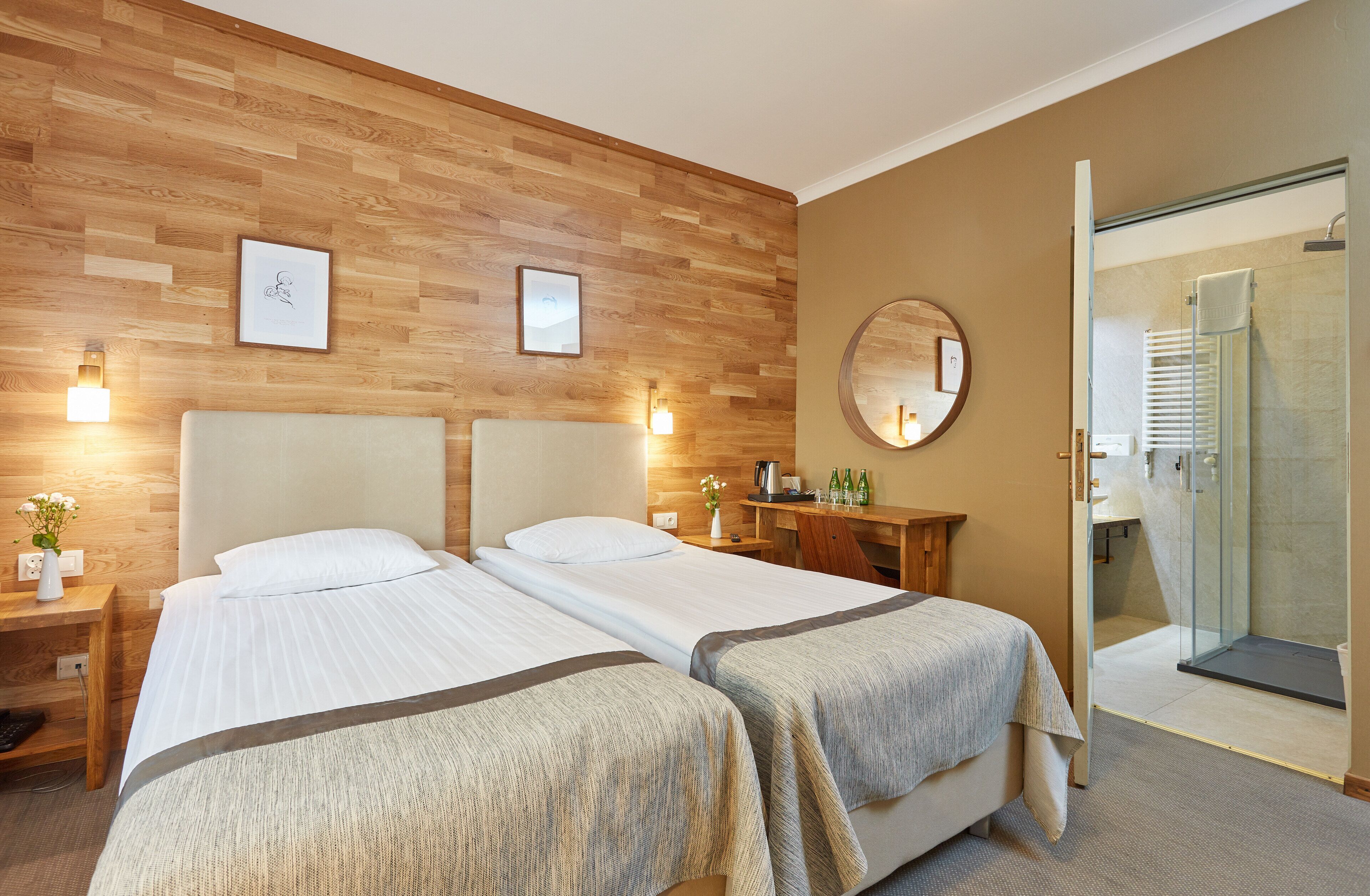 A hotel room with two beds, wooden paneling on the walls, a round mirror, and a glass-enclosed shower.