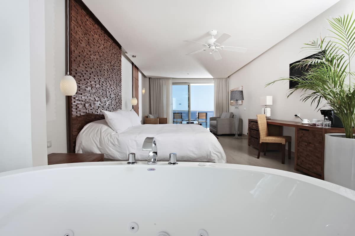 A modern hotel room with a large bed, a sitting area with a desk, a large window, and a balcony with a view.