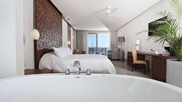 A modern hotel room with a large bed, a sitting area with a desk, a large window, and a balcony with a view.