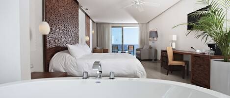 A modern hotel room with a large bed, a sitting area with a desk, a large window, and a balcony with a view.