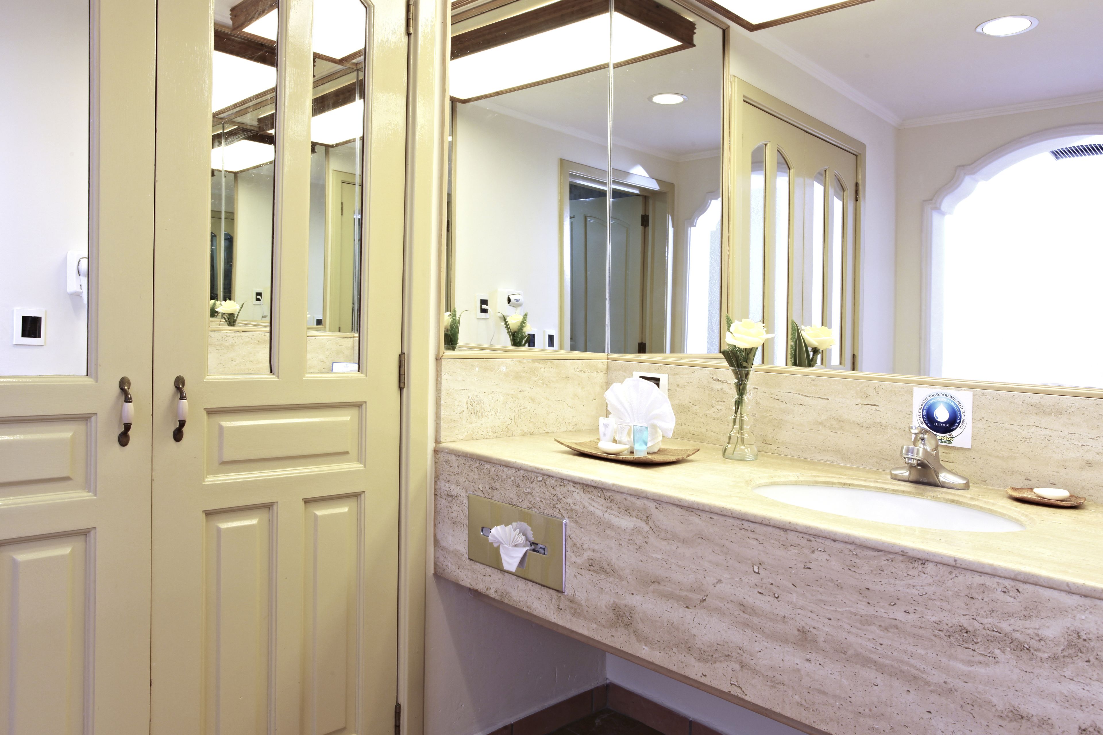Ett badrum med ett handfat i beige marmor, en stor spegel och en handduksdispenser.